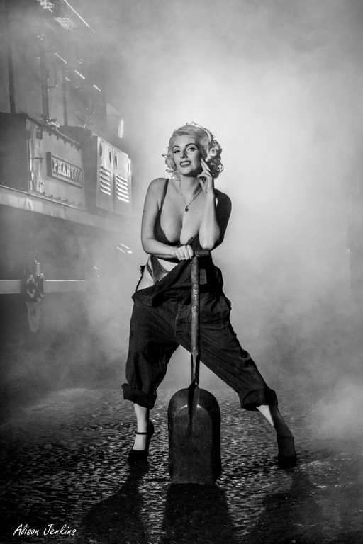 fashion model in revealing dungarees holding coal shovel next to steam train