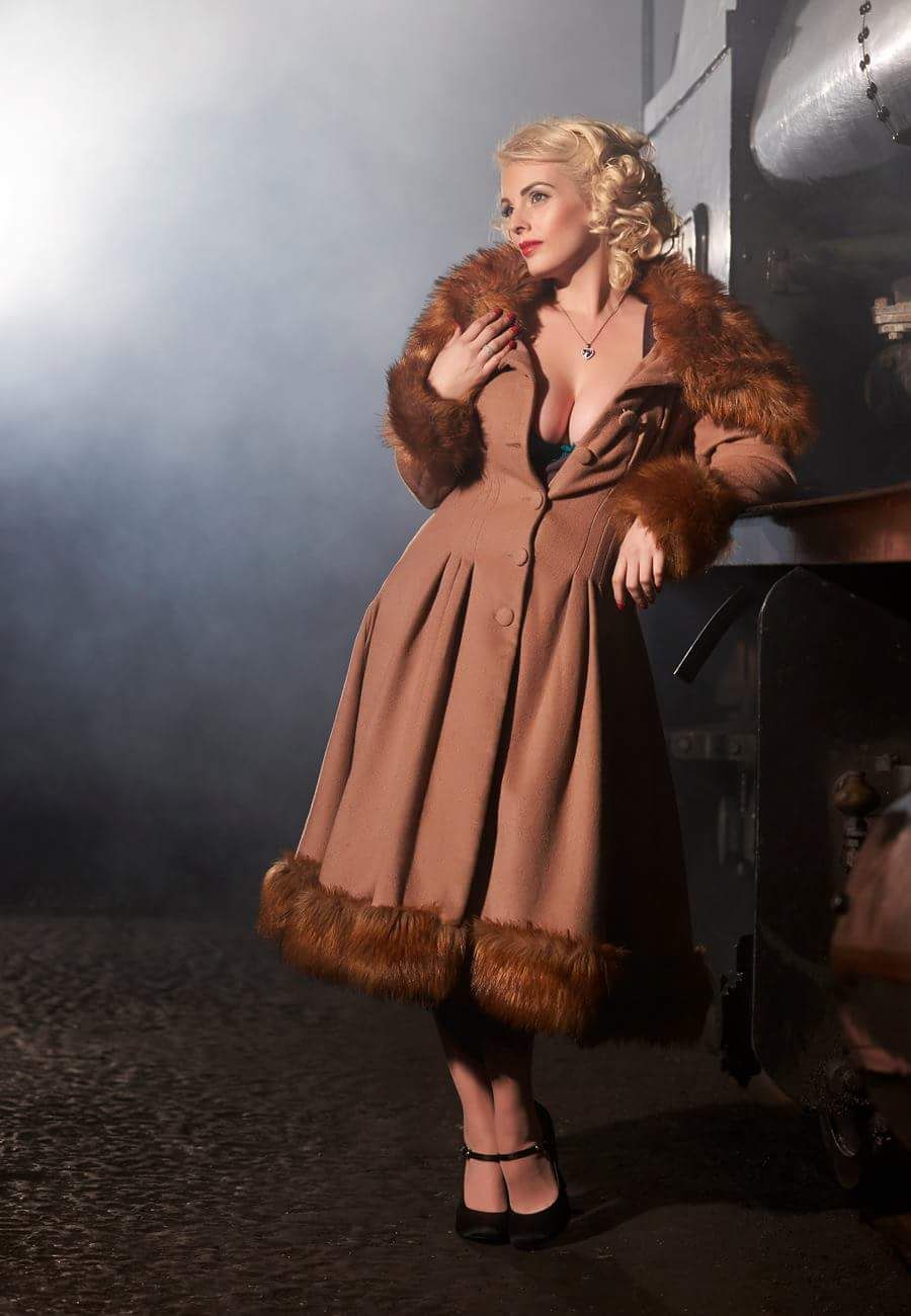 fashion model in long brown fur trimmed coat leaning on steam train